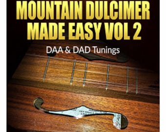 以下が含まれることがあります： 「PLAYING THE MOUNTAIN DULCIMER MADE EASY VOL 2」の文字が金色で書かれた、マウンテンダルシマーのクローズアップ。本の表紙には「DAA & DAD Tunings」と「JEFFREY A LAMBERT」の文字も含まれています。ダルシマーは木製です。