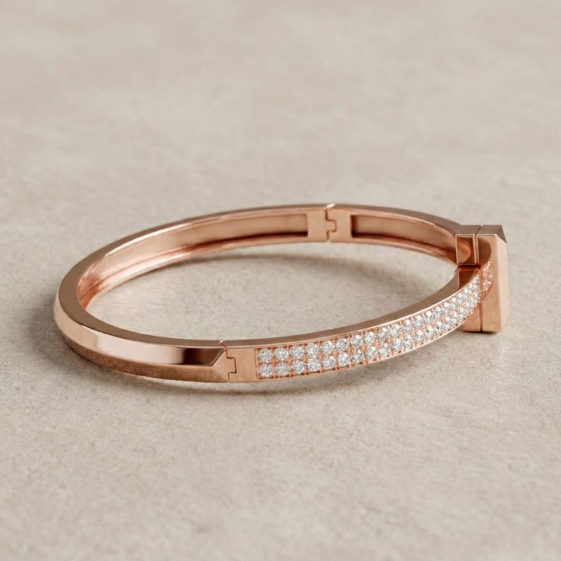 May include: Rose gold hinged bracelet with a row of small, sparkling white stones. The bracelet has a rectangular clasp and a smooth, polished finish. A piece of jewelry.