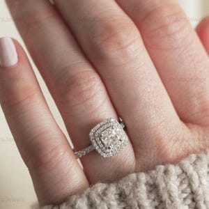 May include: A square-cut diamond ring with a double halo of smaller diamonds. The ring is set on a delicate band, and the diamonds sparkle. The ring is a piece of jewellery.
