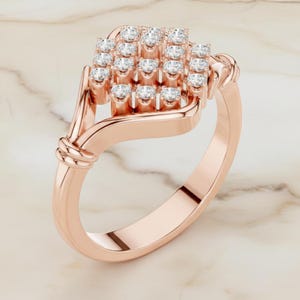 K&ouml;nnte beinhalten: Ros&eacute;goldener Ring mit einer Ansammlung kleiner, runder, klarer Edelsteine in quadratischer Form. Das Band hat eine glatte, polierte Oberfl&auml;che mit einem Knotendesign an der Seite. Der Ring wird auf einer Marmoroberfl&auml;che pr&auml;sentiert.