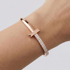 May include: Rose gold bracelet with a T-shaped design and a row of sparkling white gemstones. The bracelet is a bangle style and is worn on the wrist. The metal has a polished finish.