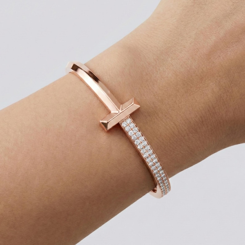 May include: Rose gold bracelet with a T-shaped design and a row of sparkling white gemstones. The bracelet is a bangle style and is worn on the wrist. The metal has a polished finish.