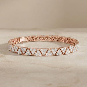 May include: Rose gold bracelet featuring a repeating triangular design. Each triangle is encrusted with small, sparkling white stones. The bracelet is displayed on a neutral, light-colored surface.