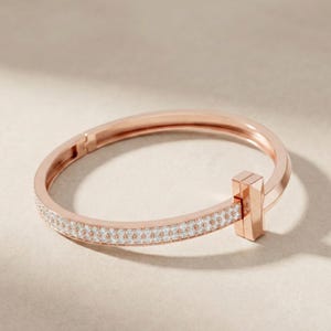 May include: Rose gold bracelet with a row of small, sparkling white stones. The bracelet has a rectangular clasp and a smooth, polished finish. The design is simple and elegant, suitable for everyday wear or special occasions.