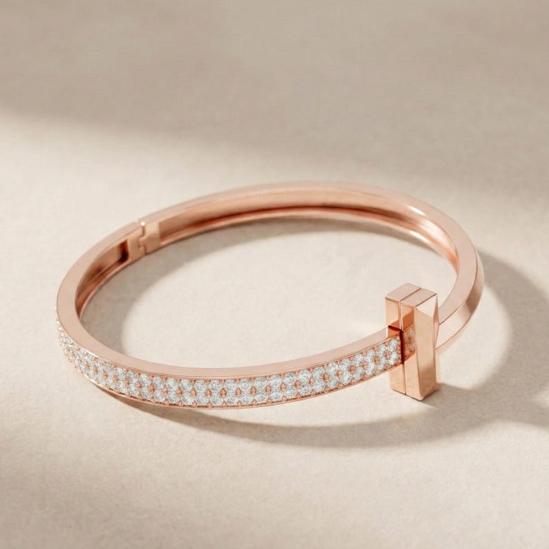 May include: Rose gold bracelet with a row of small, sparkling white stones. The bracelet has a rectangular clasp and a smooth, polished finish. The design is simple and elegant, suitable for everyday wear or special occasions.