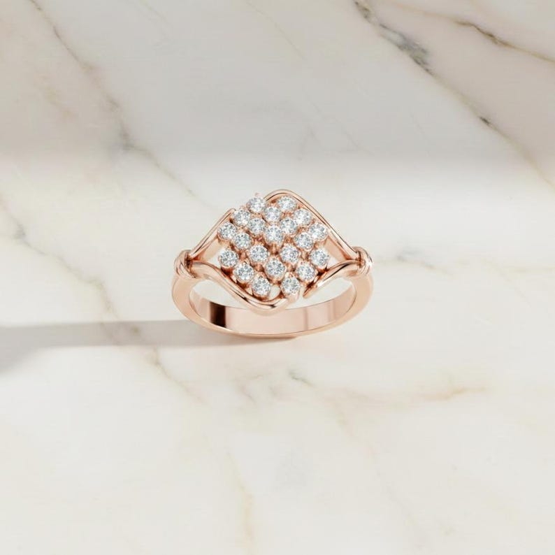 K&ouml;nnte beinhalten: Ros&eacute;goldener Ring mit einem quadratischen Cluster aus kleinen, runden, klaren Edelsteinen. Der Ring hat ein zartes Band und dekorative Akzente an den Seiten, vor einem Marmorhintergrund.
