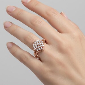 K&ouml;nnte beinhalten: Ros&eacute;goldener Ring mit einem quadratischen Cluster aus kleinen, runden, klaren Edelsteinen. Das Band des Rings hat ein gewebtes Design, und die Edelsteine sind in einem quadratischen Rahmen gefasst. Der Ring wird auf einer Hand pr&auml;sentiert.