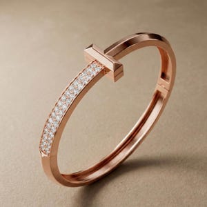 May include: Rose gold bangle bracelet with a T-shaped accent. One side of the bracelet is encrusted with small, sparkling gemstones. The bracelet has a hinged clasp for easy wear and is displayed against a neutral background.
