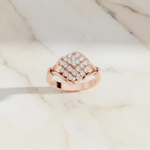 K&ouml;nnte beinhalten: Ros&eacute;goldener Ring mit einem quadratischen Cluster aus kleinen, runden Diamanten. Der Ring hat ein zartes Band und eine dekorative Fassung um das Diamantencluster. Der Ring wird auf einer wei&szlig;en Marmoroberfl&auml;che pr&auml;sentiert.