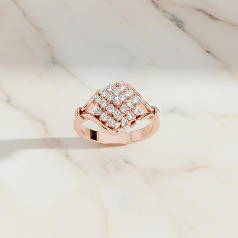 K&ouml;nnte beinhalten: Ros&eacute;goldener Ring mit einem quadratischen Cluster aus kleinen, runden Diamanten. Der Ring hat ein zartes Band und eine dekorative Fassung um das Diamantencluster. Der Ring wird auf einer wei&szlig;en Marmoroberfl&auml;che pr&auml;sentiert.