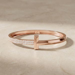May include: Rose gold bracelet with a T-shaped design. One side of the bracelet is encrusted with small, sparkling stones. The bracelet is displayed on a neutral background.