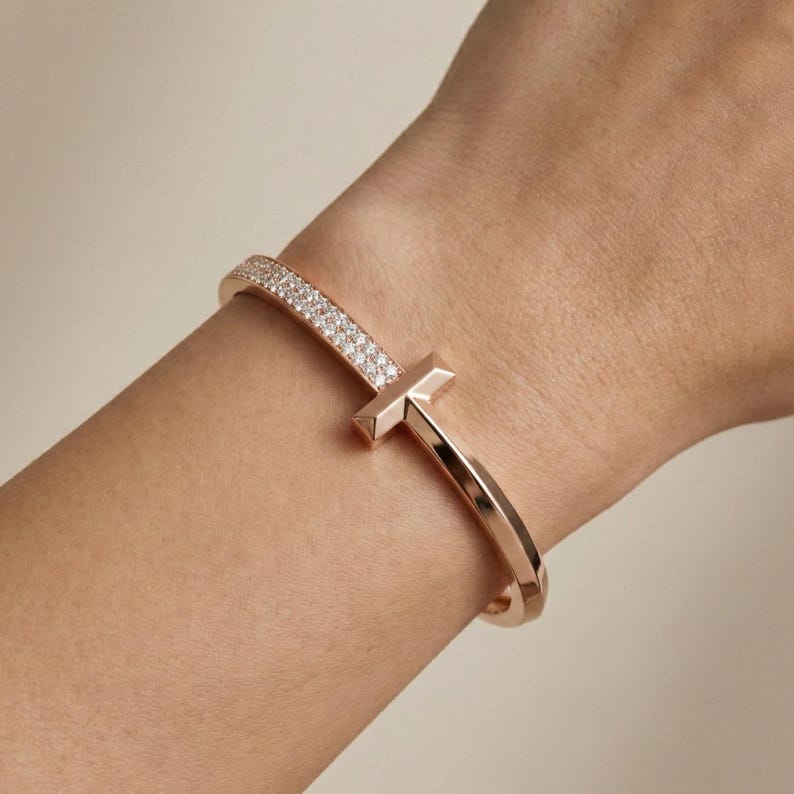 May include: Rose gold bracelet with a T-shaped design. One side of the bracelet is encrusted with small, sparkling stones. The bracelet is a cuff style, with a smooth, polished finish.