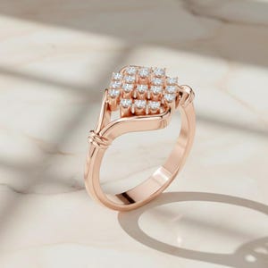 K&ouml;nnte beinhalten: Ros&eacute;goldener Ring mit einer Ansammlung kleiner, runder, klarer Edelsteine in quadratischer Form. Der Ring hat ein zartes, geschwungenes Design mit einem Knotendetail am Band. Der Ring wird auf einer Marmoroberfl&auml;che pr&auml;sentiert.