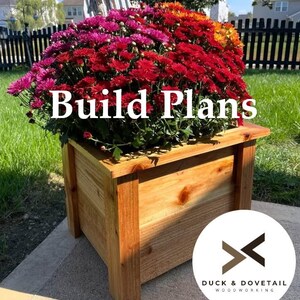 May include: A wooden planter box filled with vibrant red, pink, and orange chrysanthemums. The box is made of light-colored wood and has a simple, rectangular design. Text on the image reads "Build Plans". The Duck & Dovetail Woodworking logo is in the bottom right corner.