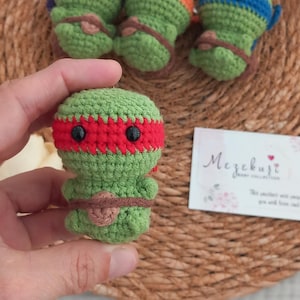 May include: Handmade green crochet keychains shaped like Teenage Mutant Ninja Turtles, each with a different colored mask: purple, orange, and blue. The keychains are on a woven background. A small card with the text "Mezebuzt" is also visible.