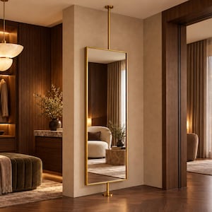 Pode incluir: Um espelho retangular do chão ao teto com uma moldura fina de metal dourado, montado entre o chão e o teto com uma haste vertical dourada. O espelho reflete um quarto com uma poltrona cor de creme e uma pequena mesa de pedra.
