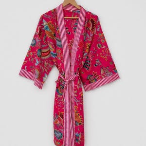 May include: A vibrant pink robe with a floral pattern and striped trim. The robe is hanging on a wooden hanger against a white background. The robe has a matching belt.