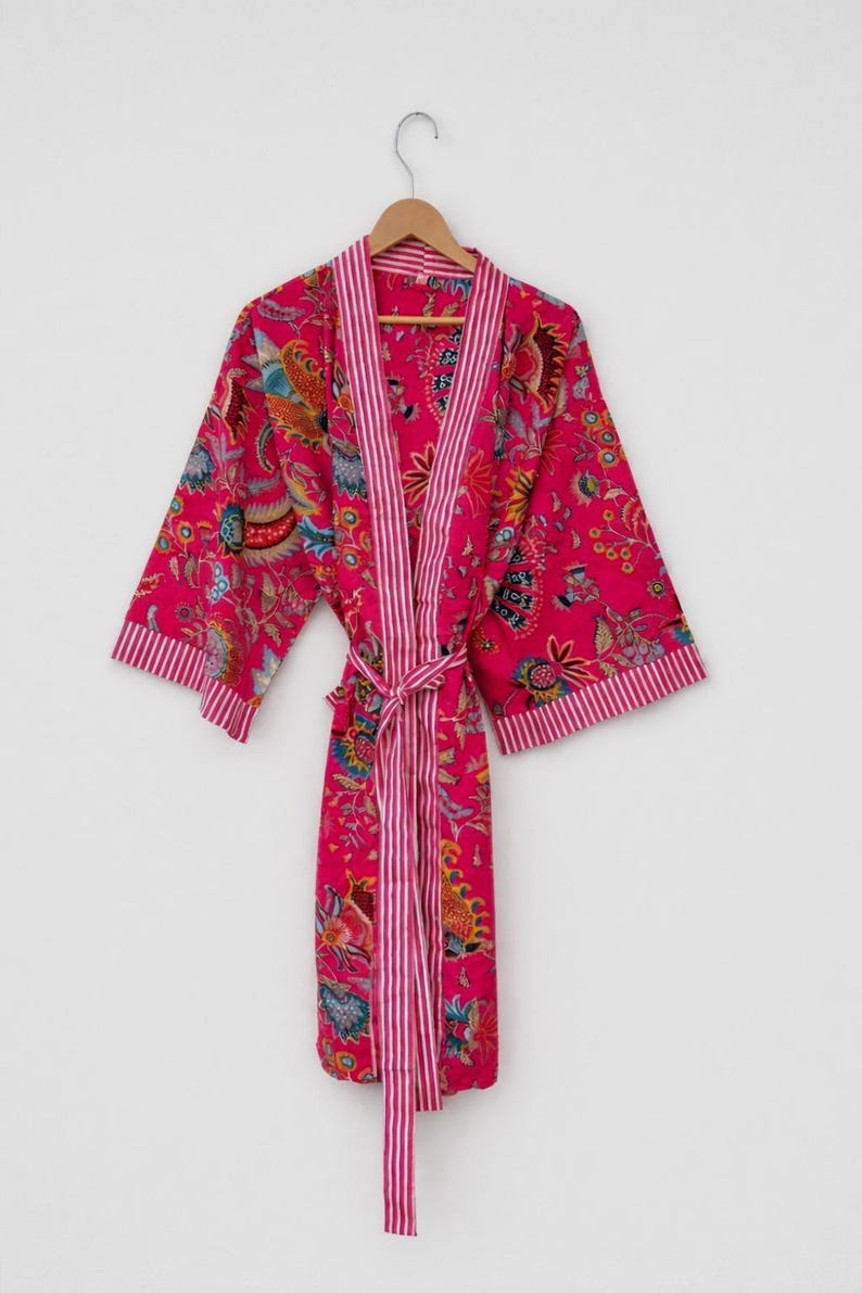 May include: A vibrant pink robe with a floral pattern and striped trim. The robe is hanging on a wooden hanger against a white background. The robe has a matching belt.