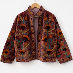 May include: A burgundy velvet jacket with floral embroidery in orange, teal, and gold. The jacket has a mandarin collar and a striped lining. The embroidery features stylized flowers and leaves, creating a bohemian aesthetic.