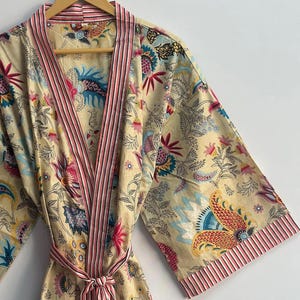 May include: A beige floral print robe with a pink striped trim and belt. The robe features a colorful botanical pattern with flowers and leaves in shades of pink, blue, and yellow. The robe is hanging on a wooden hanger.