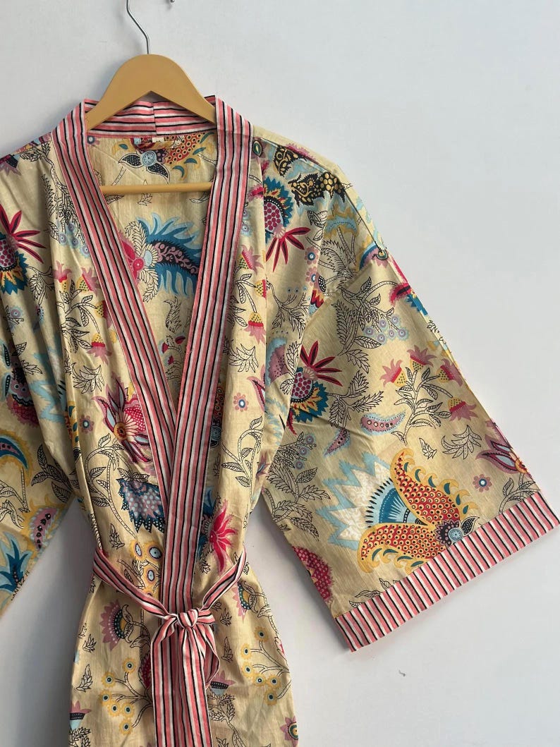May include: A beige floral print robe with a pink striped trim and belt. The robe features a colorful botanical pattern with flowers and leaves in shades of pink, blue, and yellow. The robe is hanging on a wooden hanger.
