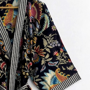 May include: A dark blue robe with a vibrant floral pattern in red, yellow, blue, and green. The robe features black and white striped trim on the edges and a matching tie belt. The sleeves are three-quarter length.