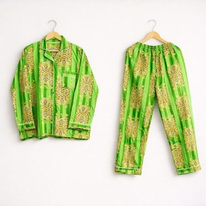 May include: A green pajama set with a tiger print. The set includes a button-down shirt and matching pants, both hanging on wooden hangers. The tiger print features yellow and black tigers on a green striped background. The pajamas have white trim.