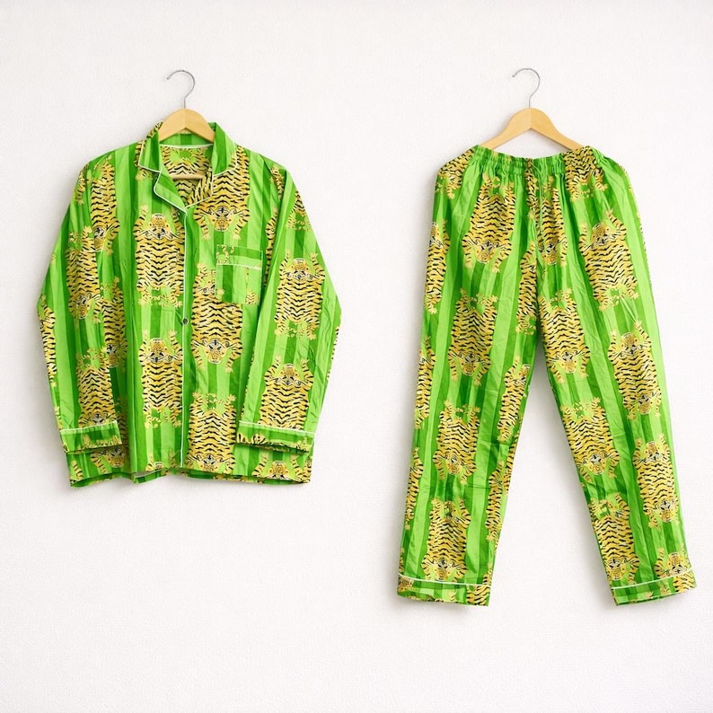 May include: A green pajama set with a tiger print. The set includes a button-down shirt and matching pants, both hanging on wooden hangers. The tiger print features yellow and black tigers on a green striped background. The pajamas have white trim.