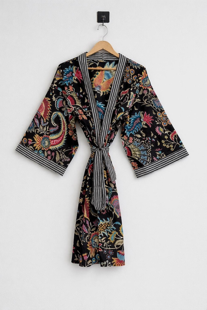 May include: A black floral print robe with a striped trim and tie. The robe features a colorful paisley and floral design in shades of red, blue, yellow, and pink. The robe is hanging on a wooden hanger.