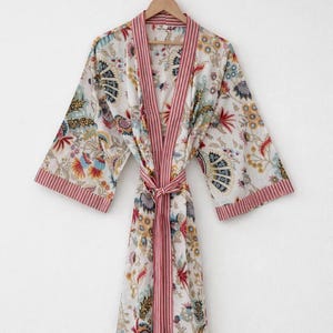 May include: A white floral print robe with a red striped trim and belt. The robe features a colorful floral pattern with blue, yellow, and red accents. The robe is hanging on a wooden hanger.