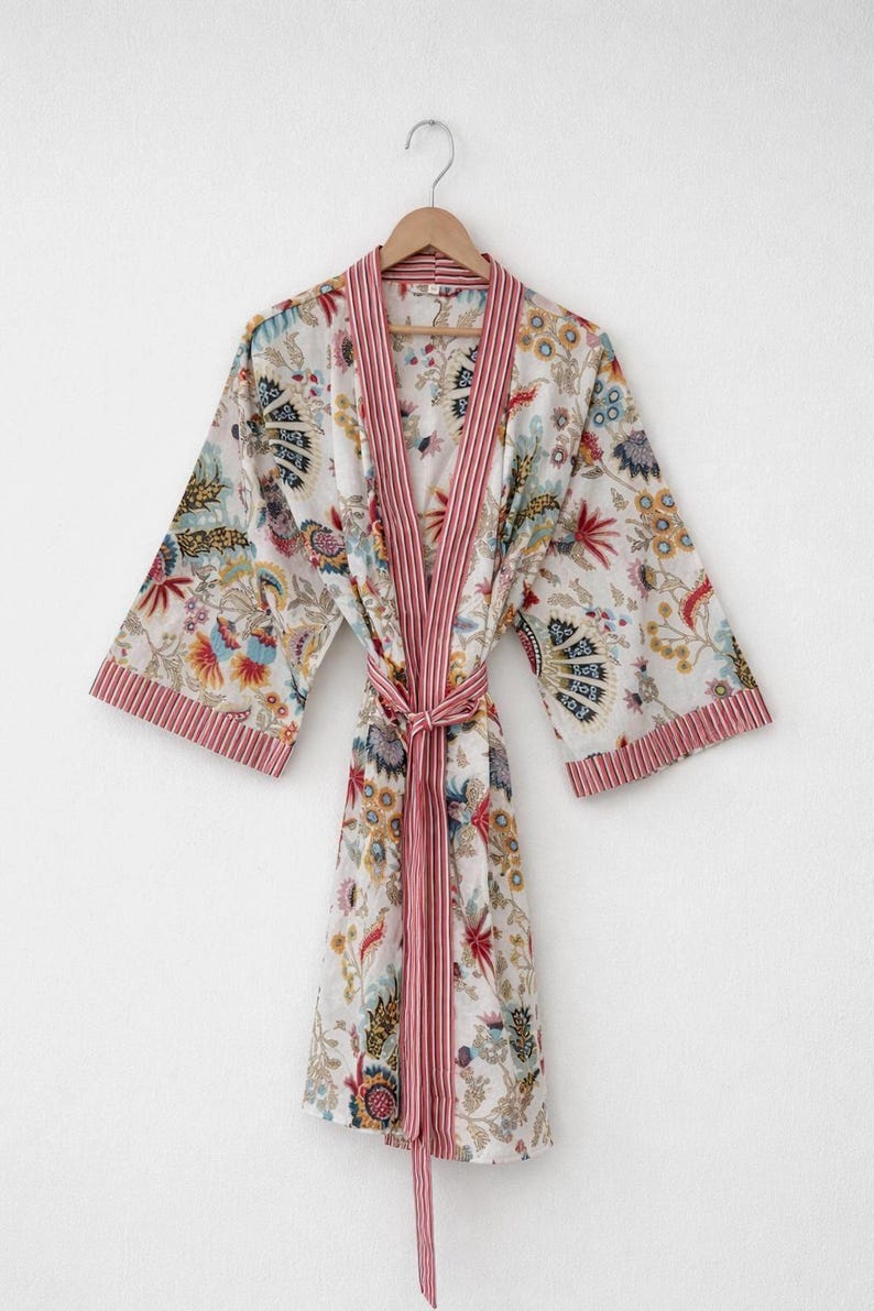 May include: A white floral print robe with a red striped trim and belt. The robe features a colorful floral pattern with blue, yellow, and red accents. The robe is hanging on a wooden hanger.