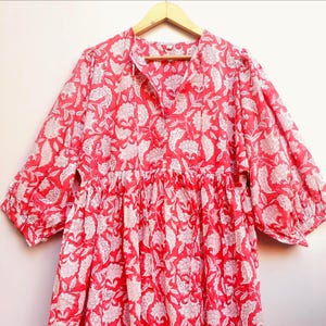 May include: A red and white floral print dress with three-quarter sleeves. The dress has a gathered waist and a V-neckline. The fabric is lightweight and appears to be cotton. The dress is hanging on a wooden hanger.
