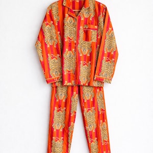 May include: Orange and red striped pajama set with a tiger print. The long-sleeved top has a button-down front and a chest pocket. The matching pants have a relaxed fit. The set is hanging on a wooden hanger.