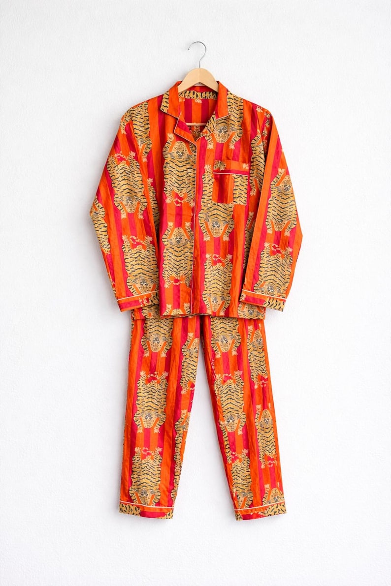 May include: Orange and red striped pajama set with a tiger print. The long-sleeved top has a button-down front and a chest pocket. The matching pants have a relaxed fit. The set is hanging on a wooden hanger.