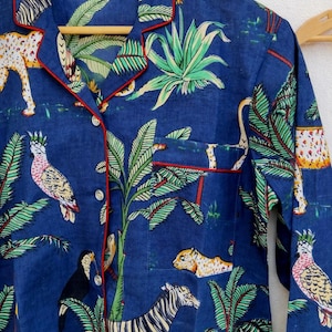 Puede incluir: Camisa de pijama azul con estampado de jungla con cebras, leopardos y loros. La camisa tiene ribetes rojos y un bolsillo en el pecho. La tela es suave y ligera.