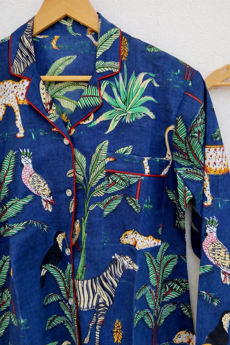Puede incluir: Camisa de pijama azul con estampado de jungla con cebras, leopardos y loros. La camisa tiene ribetes rojos y un bolsillo en el pecho. La tela es suave y ligera.