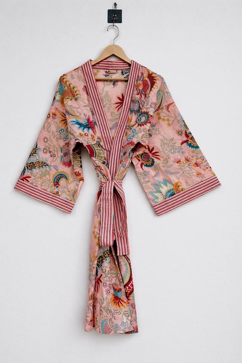 May include: A pink floral print robe with a striped trim and belt. The robe features a V-neck design and long sleeves. The floral pattern includes blue, red, and yellow accents. The robe is hanging on a wooden hanger.