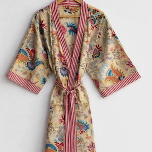 May include: A beige floral print robe with a striped pink trim and matching belt. The robe features a colorful floral pattern with blue, orange, and purple accents. It is hanging on a wooden hanger.