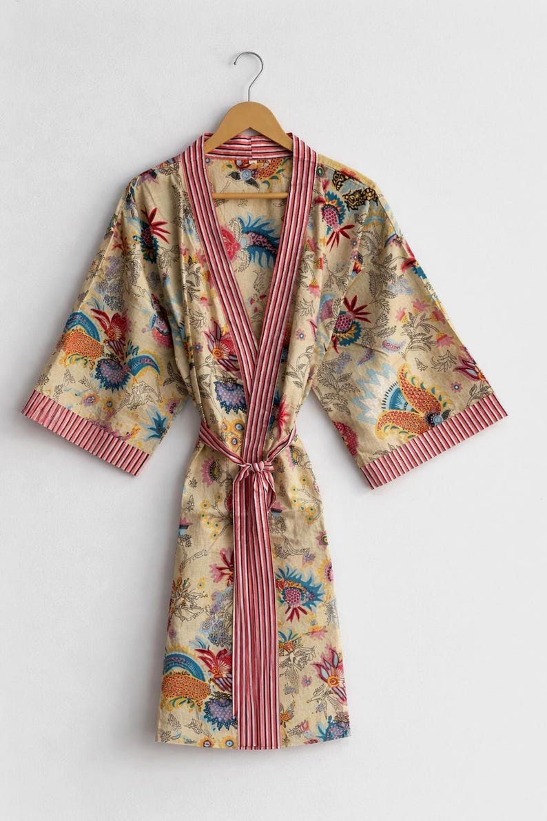 May include: A beige floral print robe with a striped pink trim and matching belt. The robe features a colorful floral pattern with blue, orange, and purple accents. It is hanging on a wooden hanger.