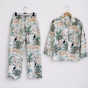Puede incluir: Conjunto de pijama blanco con estampado de tem&aacute;tica selv&aacute;tica. El conjunto incluye pantal&oacute;n largo y camisa abotonada. El estampado presenta guepardos, tucanes, palmeras y cebras. El conjunto de pijama est&aacute; colgado en una percha de madera.