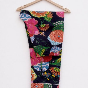 Puede incluir: Pantalones de pijama azul marino con un vibrante estampado floral en rosa, naranja, verde y azul. Los pantalones cuelgan de una percha de madera sobre un fondo blanco. El dise&ntilde;o floral incluye flores grandes y formas abstractas.