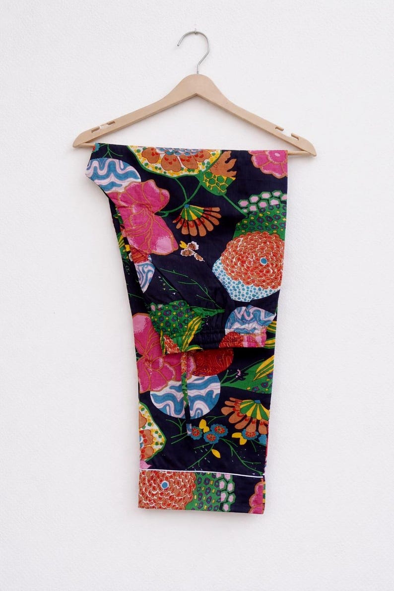Puede incluir: Pantalones de pijama azul marino con un vibrante estampado floral en rosa, naranja, verde y azul. Los pantalones cuelgan de una percha de madera sobre un fondo blanco. El dise&ntilde;o floral incluye flores grandes y formas abstractas.