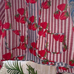 May include: A button-up jacket with a red and white striped pattern, adorned with large red strawberry illustrations. The jacket features two front pockets and is hanging on a wooden hanger. The garment has a relaxed fit.