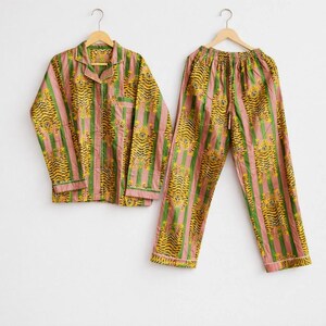 May include: A two-piece pajama set with a tiger stripe pattern. The set includes a button-down shirt and drawstring pants. The pajamas feature vertical stripes in pink, green, and yellow. The shirt has a collar and a breast pocket. The set is hanging on a wooden hanger.