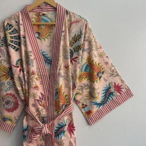May include: A light pink floral print robe with a matching belt. The robe features a colorful pattern of flowers and birds in shades of yellow, blue, and red. The trim and belt have pink and white stripes. The robe is hanging on a wooden hanger.