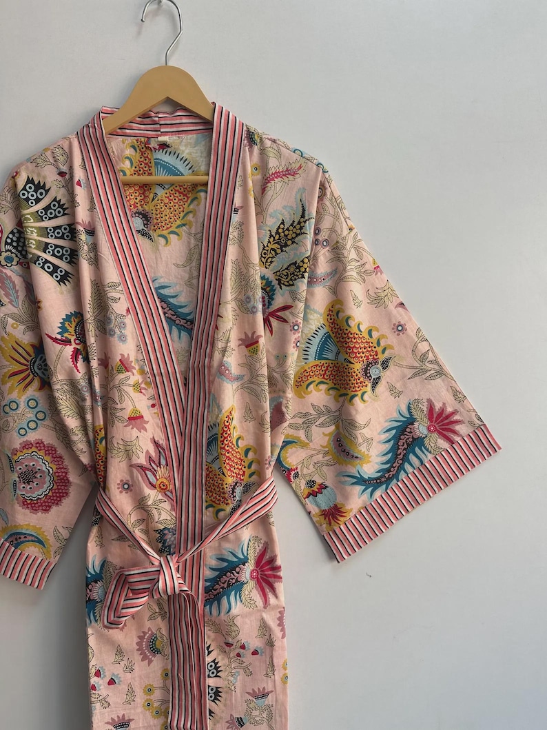 May include: A light pink floral print robe with a matching belt. The robe features a colorful pattern of flowers and birds in shades of yellow, blue, and red. The trim and belt have pink and white stripes. The robe is hanging on a wooden hanger.