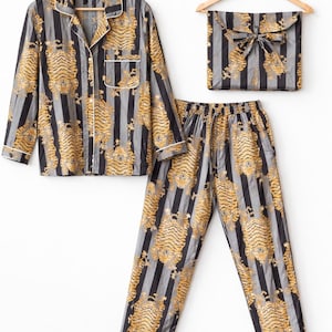 May include: A matching pajama set featuring a button-down shirt, pants, and a storage pouch. The set is patterned with vertical black and gray stripes and gold tiger illustrations. White trim accents the shirt and pants.