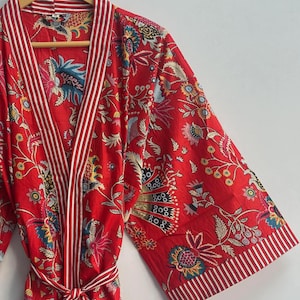 May include: A red floral print robe with white and red striped trim. The robe features a tie belt and wide sleeves. The floral pattern includes blue, yellow, and green accents. The robe is hanging on a wooden hanger.