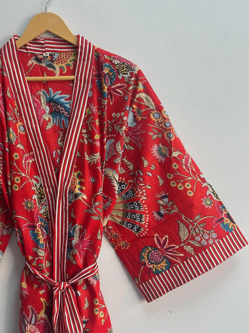 May include: A red floral print robe with white and red striped trim. The robe features a tie belt and wide sleeves. The floral pattern includes blue, yellow, and green accents. The robe is hanging on a wooden hanger.