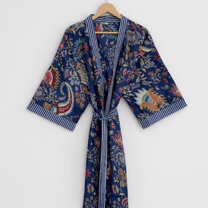 May include: A navy blue robe with a floral pattern in red, yellow, and green. The robe has white and blue striped trim on the collar and sleeves. A matching belt is tied at the waist. The robe is hanging on a wooden hanger.